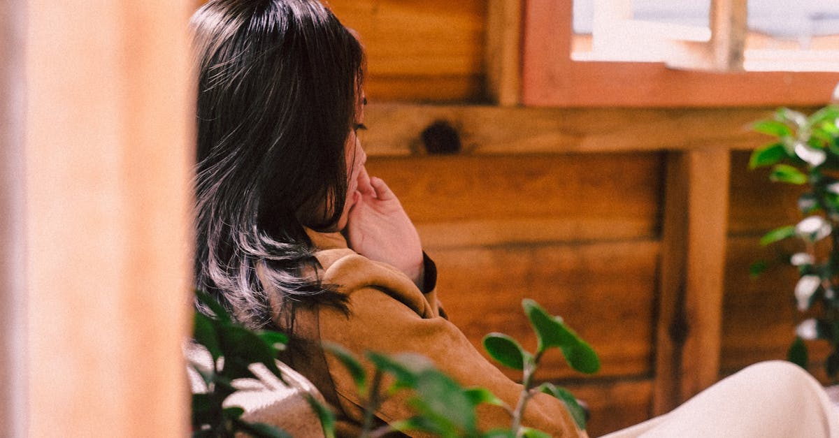 Asian woman sitting indoors in a wooden house, surrounded by greenery, reflecting tranquility.